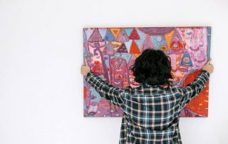 Girl hangs a painting on a white wall above a collection of cardboard boxes.